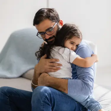 Pai abraça filha, a criança está com uma carinha triste